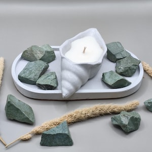 Può includere: Una candela bianca con stoppino in legno all'interno di un portacandela a forma di conchiglia su un vassoio ovale grigio. Pietre verdi circondano il vassoio, con steli di grano secco. L'insieme evoca un'estetica naturale e rilassante.