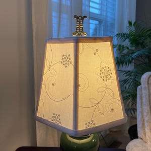 Puede incluir: Lámpara de mesa decorativa con base de cerámica verde y pantalla cuadrada. La pantalla crema presenta motivos florales bordados y un remate dorado. La lámpara está colocada sobre una mesa de madera oscura.