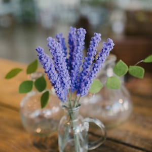May include: A small glass vase with a handle holds a bouquet of purple flowers. Two other glass vases are in the background, along with green leaves. The arrangement sits on a wooden table.