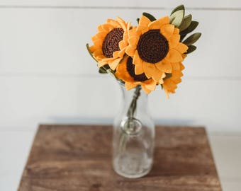 May include: A bouquet of handcrafted felt sunflowers featuring bright yellow petals and dark brown centers, accompanied by green felt leaves. The flowers are arranged in a clear glass bottle with a narrow neck, showcasing the stems inside the vessel.