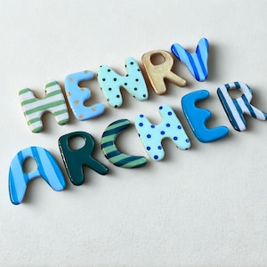 May include: A set of decorative wooden letters spelling out the name HENRY ARCHER. Each letter features a unique pattern in shades of blue, green, and gold, including stripes, polka dots, and solid colors, arranged in two rows on a light surface.