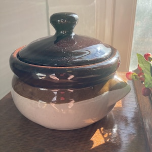 May include: A round ceramic lidded pot featuring a two-tone glaze with a dark brown top and a speckled cream-colored base. The lid has a central knob handle, and the pot includes a small integrated pouring spout on the side.