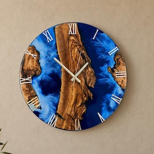 Könnte beinhalten: Eine kreisförmige Wanduhr mit einem zentralen vertikalen Stück natürlicher Holzmaserung, das von tiefblauem Harz umgeben ist. Das Zifferblatt zeigt weiße römische Ziffern und dünne, helle Metallzeiger, die einen Kontrast zwischen dem organischen Holz und der glatten, leuchtend blauen Harzoberfläche bilden.