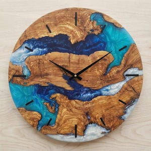 May include: Circular wooden wall clock with a distinctive design. The clock face showcases natural wood grain patterns with blue and white resin accents, and black clock hands. The clock measures approximately 30 cm in diameter.