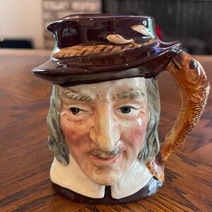 May include: A ceramic mug featuring a man's face with a hat and a fish-shaped handle. The mug is primarily brown with accents of tan, white, and pink. The mug has a detailed design and is likely a collectible item.