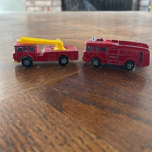 May include: Two vintage red toy fire trucks. One has a yellow extending ladder, the other has a detailed cab. Both have black wheels and are set on a wooden surface. These collectible die-cast metal vehicles are likely from the 1970s or 1980s.