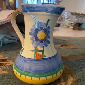 May include: A ceramic pitcher with a floral design. The pitcher features a blue and yellow flower on a cream-colored background. The base is yellow with green and blue stripes, and the handle has orange accents.