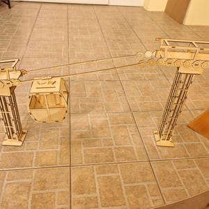 Op de afbeelding: Een houten model van een kabelbaan, met een gondel die tussen twee steuntorens hangt. De torens hebben een tralieontwerp en de gondel heeft een rechthoekige vorm. Het model is gemaakt van lichtgekleurd hout.