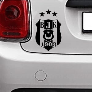 May include: Black and white emblem of a soccer team, featuring a shield with the letters BJK, a crescent moon and star, and the year 1903. The emblem is affixed to the rear of a white vehicle.