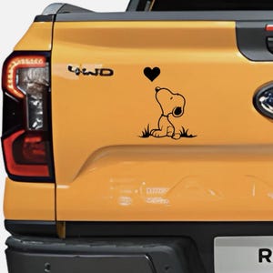 Könnte beinhalten: Schwarzer Vinyl-Aufkleber von Snoopy, der auf ein schwarzes Herz blickt. Der Aufkleber befindet sich auf der Rückseite eines gelben Lastwagens. Der Lastwagen hat eine schwarze Stoßstange und ein rotes Rücklicht.