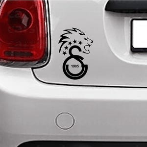 Puede incluir: Calcoman&iacute;a negra para coche con la cabeza de un le&oacute;n, estrellas y las letras "GS" con el n&uacute;mero "1905" en un coche blanco. El le&oacute;n tiene una melena detallada y mira hacia la derecha.