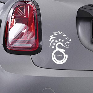 Puede incluir: Calcoman&iacute;a blanca de una cabeza de le&oacute;n rugiente, estrellas y las letras "GS" con el n&uacute;mero "1905" en un coche gris. La calcoman&iacute;a est&aacute; en la parte trasera del coche. El piloto trasero del coche es visible.