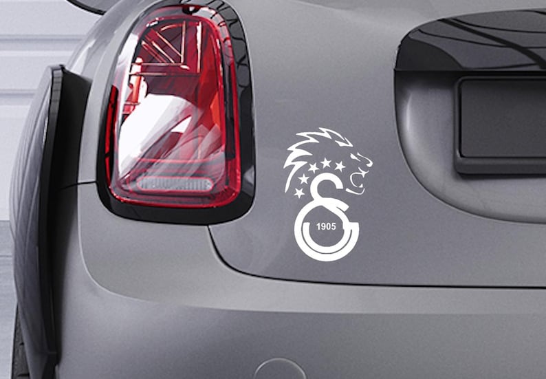 Puede incluir: Calcoman&iacute;a blanca de una cabeza de le&oacute;n rugiente, estrellas y las letras "GS" con el n&uacute;mero "1905" en un coche gris. La calcoman&iacute;a est&aacute; en la parte trasera del coche. El piloto trasero del coche es visible.