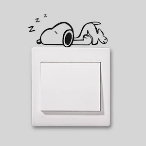 Op de afbeelding: Een witte lichtschakelaar met een Snoopy-sticker die erboven slaapt. De zwart-witte sticker toont Snoopy die ligt met het woord 'z' boven zijn hoofd. De lichtschakelaar is rechthoekig en heeft een wit frame.