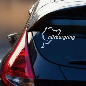 Puede incluir: Adhesivo de vinilo blanco con el contorno del circuito de Nürburgring y texto, aplicado en la luneta trasera de un coche. El adhesivo es una representación estilizada del famoso circuito alemán, con la palabra "nürburgring" impresa debajo.
