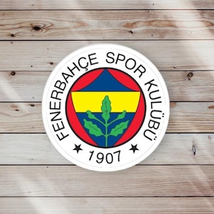 May include: A round white emblem with the text "FENERBAHÇE SPOR KULÜBÜ" in black around the edge. The emblem features a yellow and blue design with a green leaf and the year "1907". The emblem is set against a wooden background.