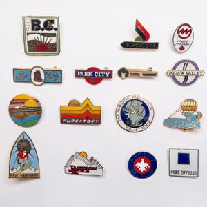 May include: A collection of fourteen metal enamel pins featuring various ski resort logos and mountain themes. The pins display colorful designs including text such as Ski B.C., Blackcomb, Whistler Mountain, Park City, Deer Valley, Squaw Valley, Lake Tahoe, Purgatory, Mammoth Mountain, Keystone, Mt. Sunapee, Jackson Hole, Phoenix Ski Club, and a blue square labeled More Difficult.