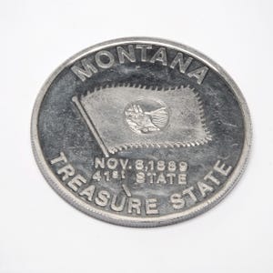 May include: A circular silver-toned commemorative coin featuring the text MONTANA at the top and TREASURE STATE at the bottom. The center displays a flag with a circular emblem, with the text NOV. 8, 1889 and 41st STATE printed below it.