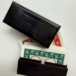 May include: A black playing card case with a matching lid, partially open to reveal a deck of cards. The cards have a white background with green and red decorative patterns.