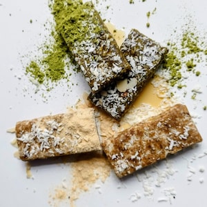 May include: An overhead shot shows three energy bars, each with different toppings, scattered on a white surface. One bar is covered in green powder, another with coconut flakes, and the third with a tan powder.