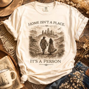 May include: A cream-colored short-sleeve t-shirt featuring a dark brown graphic print of two people walking toward a stone circle in a landscape. The text reads, "HOME ISN'T A PLACE, IT'S A PERSON." The shirt has a crew neckline and a relaxed fit.