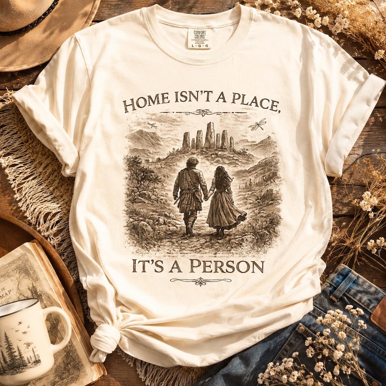 May include: A cream-colored short-sleeve t-shirt featuring a dark brown graphic print of two people walking toward a stone circle in a landscape. The text reads, "HOME ISN'T A PLACE, IT'S A PERSON." The shirt has a crew neckline and a relaxed fit.
