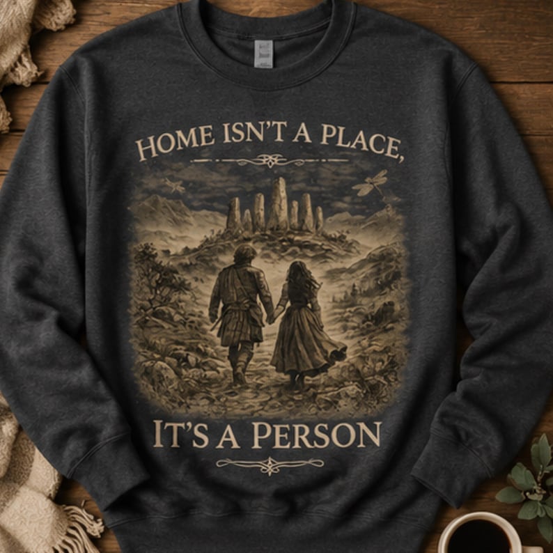 May include: A dark charcoal gray crewneck sweatshirt featuring a sepia-toned graphic of two people walking toward a stone circle. The text "HOME ISN'T A PLACE, IT'S A PERSON" is printed in a light beige font above and below the central illustration.
