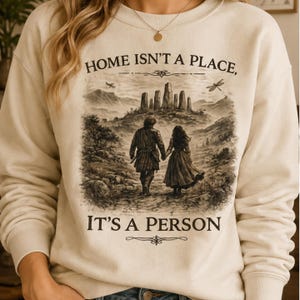 May include: A cream-colored long-sleeve sweatshirt featuring a dark brown graphic print of two figures walking toward stone pillars. The text reads, "HOME ISN'T A PLACE, IT'S A PERSON." The design includes a dragonfly and a bird in the sky.