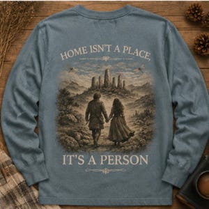 May include: A dusty blue long-sleeve shirt featuring a graphic print on the back. The illustration depicts two figures walking toward a stone monument in a landscape. The text reads: HOME ISN'T A PLACE, IT'S A PERSON, with decorative flourishes above and below.