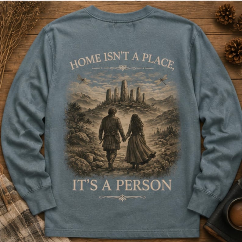 May include: A dusty blue long-sleeve shirt featuring a graphic print on the back. The illustration depicts two figures walking toward a stone monument in a landscape. The text reads: HOME ISN'T A PLACE, IT'S A PERSON, with decorative flourishes above and below.