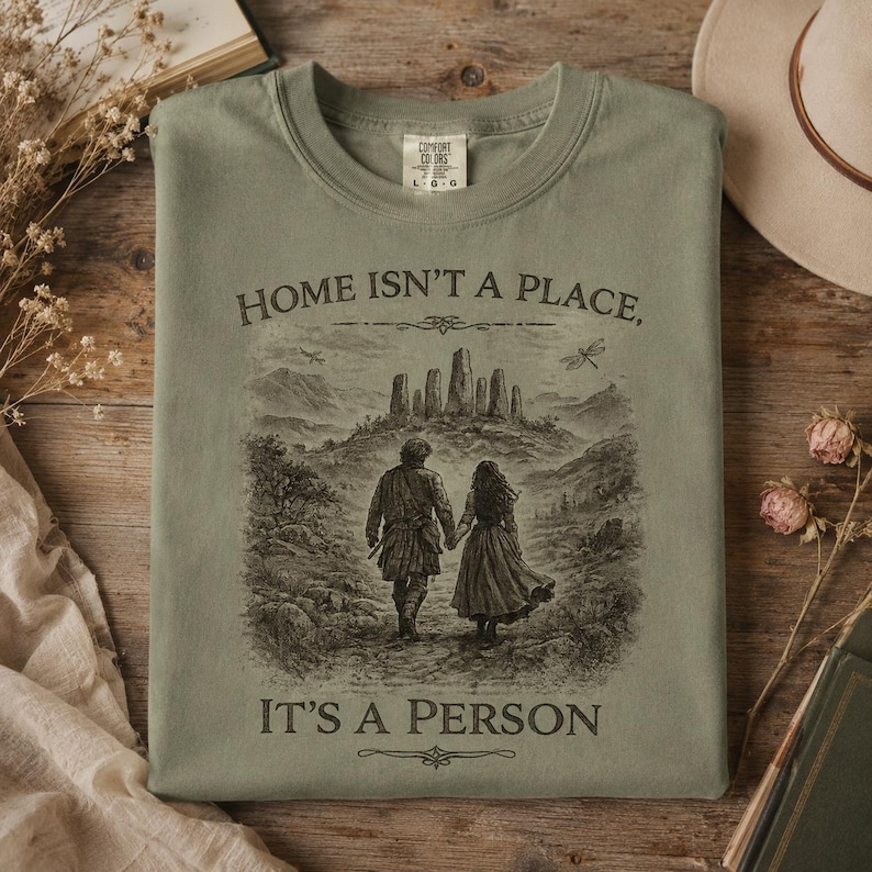 May include: A sage green crewneck t-shirt featuring a dark brown graphic of two people walking toward a stone circle. The text reads, "HOME ISN'T A PLACE, IT'S A PERSON." The shirt has a Comfort Colors label at the inner neckline indicating a size large.