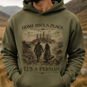 May include: An olive green hooded sweatshirt featuring a graphic print of two figures walking hand-in-hand toward a stone circle. The text "HOME ISN'T A PLACE, IT'S A PERSON" is printed in dark brown lettering above and below the central illustration.