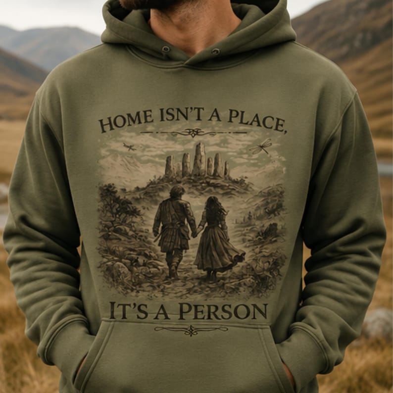 May include: An olive green hooded sweatshirt featuring a graphic print of two figures walking hand-in-hand toward a stone circle. The text "HOME ISN'T A PLACE, IT'S A PERSON" is printed in dark brown lettering above and below the central illustration.
