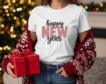 May include: White t-shirt with the text "happy NEW year" in black and pink glitter lettering. The person is wearing a red cardigan with a white snowflake pattern and holding a red gift box. The background includes a Christmas tree with lights.