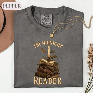 May include: A charcoal gray crew neck t-shirt featuring a graphic of a stack of books, a lit candle, a moth, and ivy. The text reads THE MIDNIGHT READER. The shirt has a Comfort Colors label in the neck, indicating size XL, TG, EG. A gold star necklace is placed on the shirt.