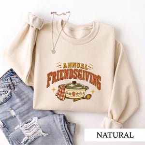 May include: A cream-colored crewneck sweatshirt featuring the text ANNUAL FRIENDSGIVING in orange and brown letters, with a graphic of a green lidded pot, a red and white checkered cloth, and a wooden spoon, paired with light blue distressed jeans.