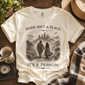 May include: A cream-colored short-sleeve t-shirt featuring a dark brown graphic of two people walking toward a stone circle. The text reads, "HOME ISN'T A PLACE, IT'S A PERSON." The shirt has a crew neckline and a Comfort Colors brand label at the neck.