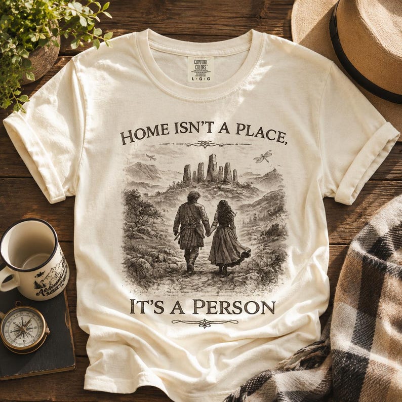 May include: A cream-colored short-sleeve t-shirt featuring a dark brown graphic of two people walking toward a stone circle. The text reads, "HOME ISN'T A PLACE, IT'S A PERSON." The shirt has a crew neckline and a Comfort Colors brand label at the neck.