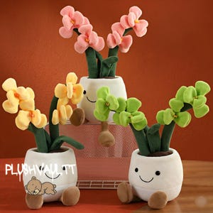 Puede incluir: Plantas de orquídeas de peluche en macetas blancas con caras sonrientes y pies marrones. Las orquídeas tienen flores rosas, amarillas y verdes. El texto "PLUSHVAULTT" es visible en una de las macetas.