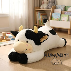 May include: A large, plush cow toy in a lying position. The toy is mainly white with black spots and accents on the ears, horns, and hooves. It has a light tan face with a pink blush and a smiling expression. The toy is on a wooden floor.