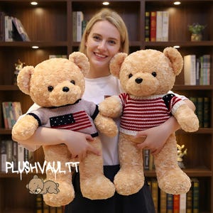 Puede incluir: Dos osos de peluche de pelaje marrón claro son sostenidos por una persona. Uno de los osos lleva un suéter con un diseño de rayas rojas y blancas, y el otro lleva un suéter con un diseño de la bandera estadounidense. Los osos tienen ojos negros y sonríen.