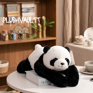 Op de afbeelding: Een zwart-witte pluche panda ligt op een witte tafel. De panda heeft zwarte vlekken rond zijn ogen, oren en ledematen. De tekst "PLUSHVAULTTT" is zichtbaar op de achtergrond.