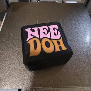 Op de afbeelding: Een zwart vierkant object met een getextureerd oppervlak voorzien van de tekst "NEE DOH" in een gestileerd lettertype. Het woord "NEE" is gedrukt in lichtroze en het woord "DOH" is gedrukt in een goudoranje kleur op de bovenzijde.