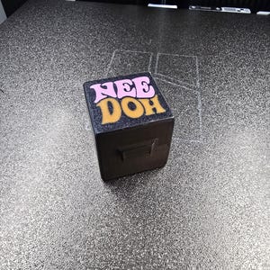 May include: A black cube-shaped object with a textured surface. The top face features the text "NEE DOH" printed in a stylised font, with "NEE" in light pink and "DOH" in a golden yellow colour. The object has a small rectangular protrusion on the side.