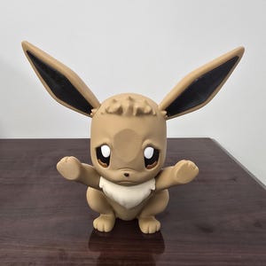 May include: A tan Eevee figurine with large ears, black tips, and a white neck ruff. The figurine has large, expressive eyes and outstretched arms. The figure is sitting on a dark brown surface.