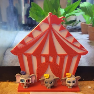 May include: A red and white circus tent with three Dumbo figurines. The tent has a red flag and red and white striped panels. The Dumbo figurines are grey with different coloured hats and accessories.