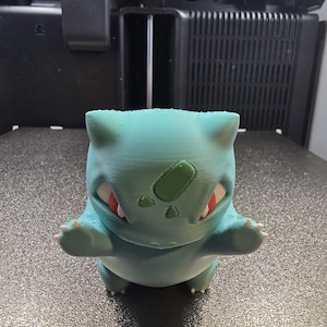 Puede incluir: Figura de Bulbasaur turquesa impresa en 3D, con ojos rojos y un bulbo verde en la espalda. La figura está sobre una superficie texturizada, con una impresora 3D en el fondo.