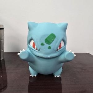 Puede incluir: Figura de Bulbasaur azul claro con ojos rojos y un bulbo verde en la espalda. La figura tiene pequeñas garras blancas y posa con los brazos extendidos. La figura está sobre una superficie marrón oscuro.
