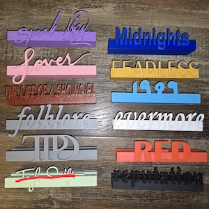 May include: A collection of colorful 3D-printed signs, each displaying a different album title in stylized lettering. The signs are in various colors, including purple, pink, blue, yellow, and red. Titles include "Speak Now," "Lover," and "Midnights."
