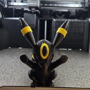 May include: A 3D printed black and yellow Umbreon figurine with pointed ears, a yellow ring on its forehead, and yellow bands on its ears. The figure is positioned on a textured print bed with the text 320 x 340 mm³ visible on the front edge.